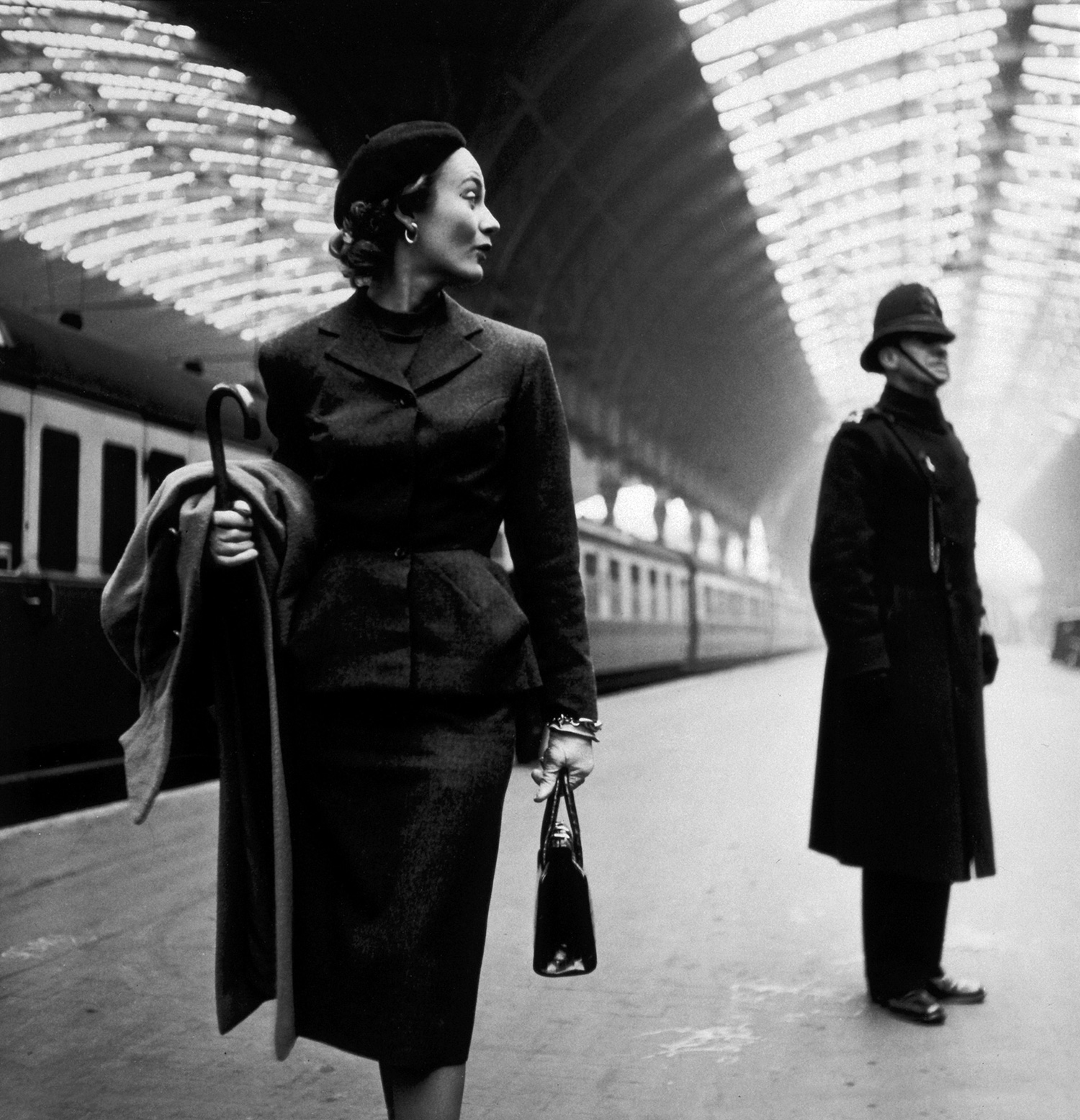 Välklädd vacker kvinna går längs en tågperrong, svartvitt fotografi. Paddington station i London. I bakgrunden en bobby med hatt
